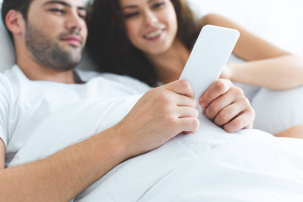 close-up view of smiling young couple using smartphone while lying together in bed - Valokuva, kuva