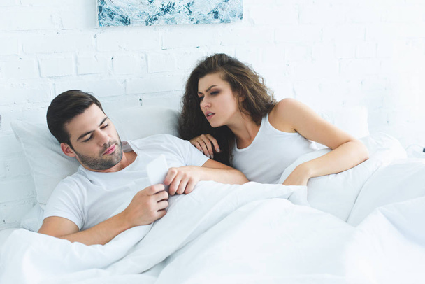 emotionale junge Frau schaut Freund mit Smartphone im Bett an, Konzept für Beziehungsschwierigkeiten - Foto, Bild