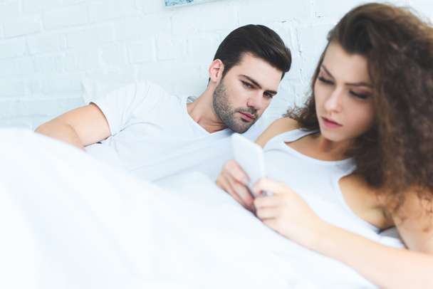 молодой человек смотрит на подругу, использующую смартфон в постели, концепция трудностей в отношениях
 - Фото, изображение