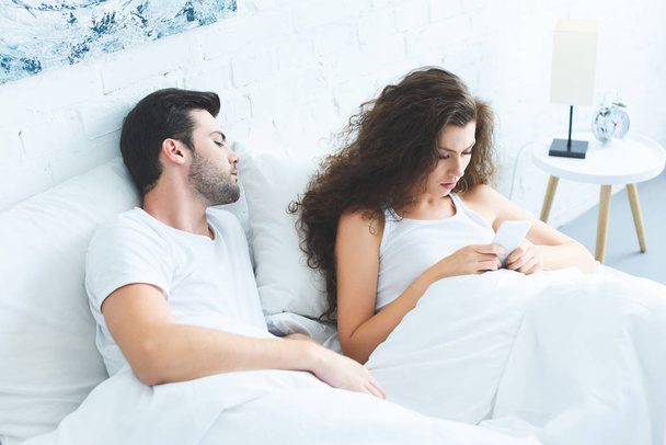 high angle view of serious young woman using smartphone while boyfriend sleeping in bed - Фото, изображение