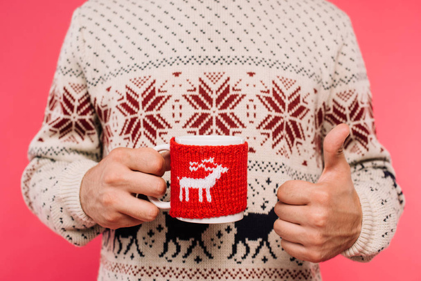 cropped image of man in sweater holding cup with deer silhouette and showing thumb up isolated on pink - Foto, Imagen