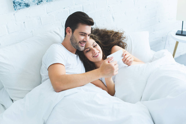 Blick aus der Vogelperspektive auf glückliches junges Paar, das gemeinsam Smartphone im Bett benutzt   - Foto, Bild