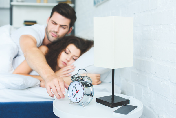 joven alcanzando el despertador mientras duerme con su novia en la cama
  - Foto, Imagen