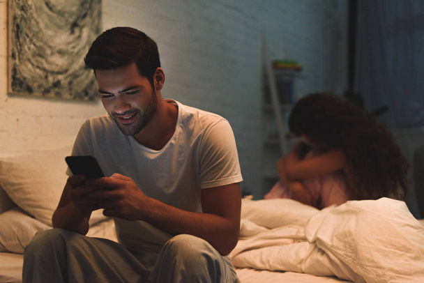uomo utilizzando smartphone mentre sconvolto fidanzata seduta sul letto di notte, concetto di difficoltà di relazione
  - Foto, immagini