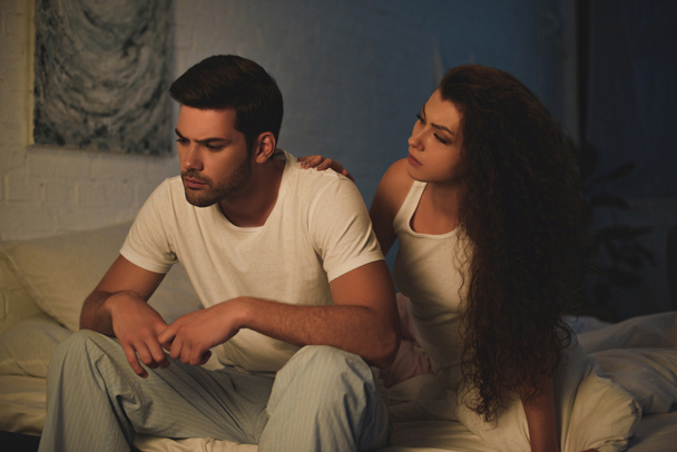 young woman looking at upset boyfriend in pajamas sitting on bed at night, relationship difficulties concept - Foto, Imagen
