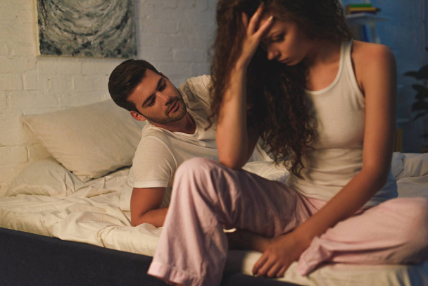 upset young woman sitting on bed while boyfriend lying behind, relationship difficulties concept - 写真・画像