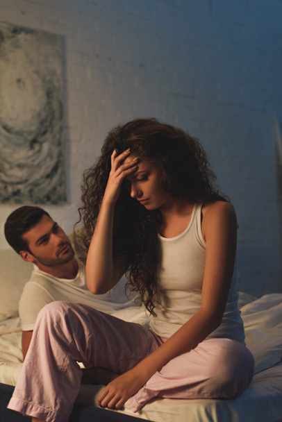 jonge man kijkend naar ontevreden vriendin zittend op bed's nachts, relatie problemen concept - Foto, afbeelding