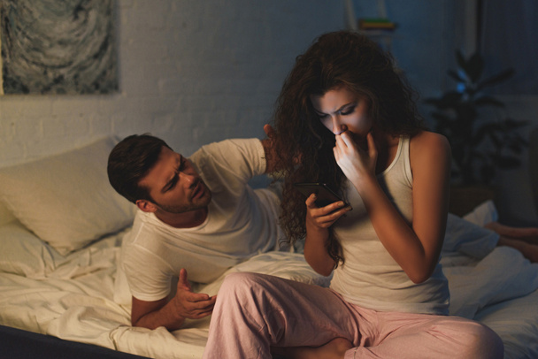Wütender junger Mann sieht aufgebrachte Freundin nachts mit Smartphone an - Foto, Bild