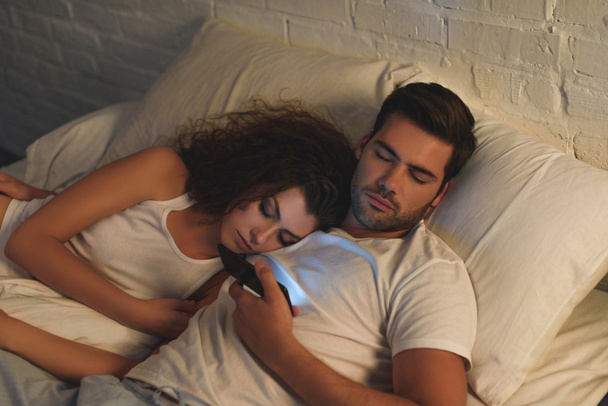 high angle view of young couple sleeping in bed, man holding smartphone - Fotografie, Obrázek
