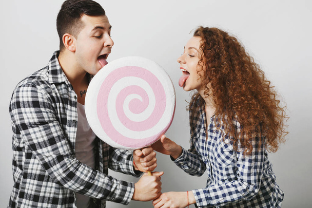 Two female and male friends lick big lollipop together, like sweets and candies, being sweety tooth, isolated over white studio background. Pretty curly woman and her boyfriend with sweet candy - Fotografie, Obrázek