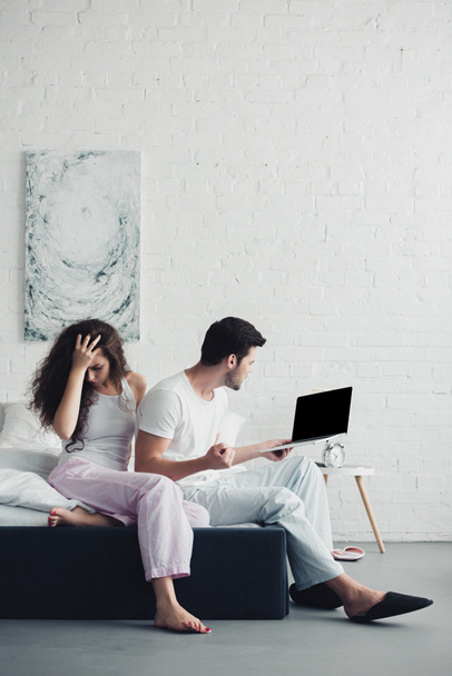 aufgebrachte junge Frau sitzt im Bett, während wütender Mann Laptop mit leerem Bildschirm benutzt, Konzept für Beziehungsschwierigkeiten - Foto, Bild