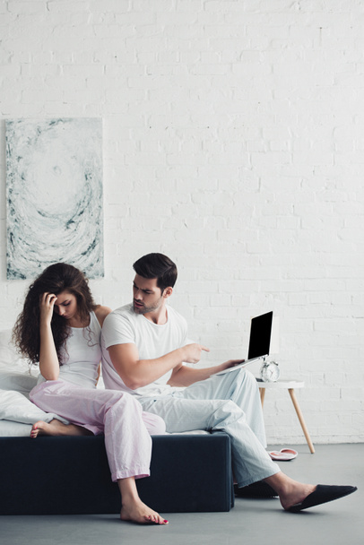 aufgebrachte junge Frau sitzt auf Bett, während Mann mit Finger auf Laptop mit leerem Bildschirm zeigt  - Foto, Bild