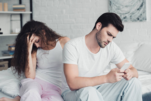 Junger Mann nutzt Smartphone, während verärgerte Frau im Bett sitzt, Beziehungsschwierigkeiten - Foto, Bild