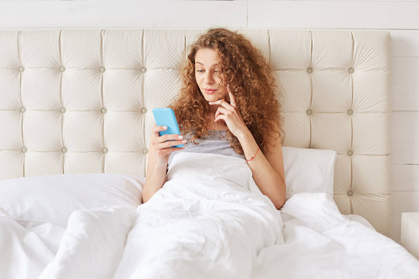 Positive attractive female watches interesting movie or chats in internet, uses modern cell phone, connected to wireless internet, enjoys leisure at bedroom on comfortable bed. People and rest concept - Foto, imagen
