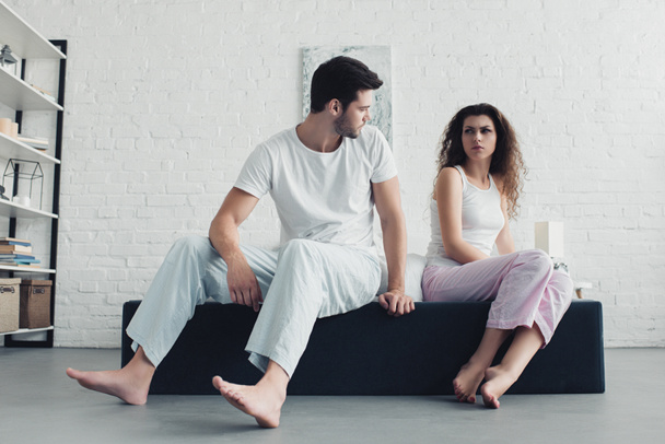 upset young couple in pajamas sitting on bed and looking at each other, relationship difficulties concept - Photo, Image