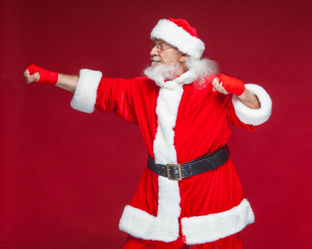 Christmas. Santa Claus with red bandages wound on his hands for boxing imitates kicks. Kickboxing, karate, boxing. Isolated on red background. - Foto, Imagem