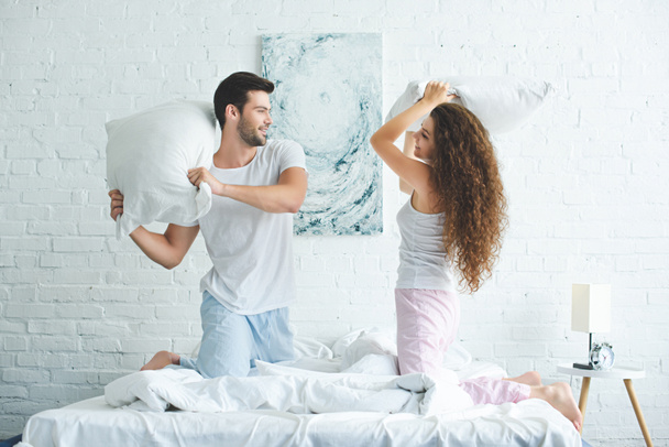 side view of happy young couple in pajamas fighting with pillows on bed   - Photo, Image
