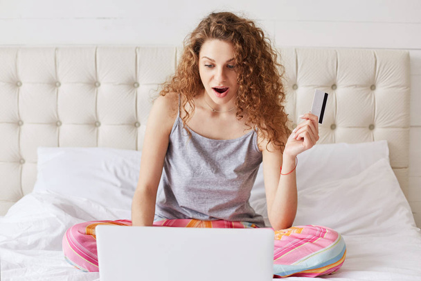 Excited female with curly har, dressed in pyjamas, uses laptop computer for online shopping, holds credit card, being shocked to realise that she has no money on her account. Online shopping - Zdjęcie, obraz