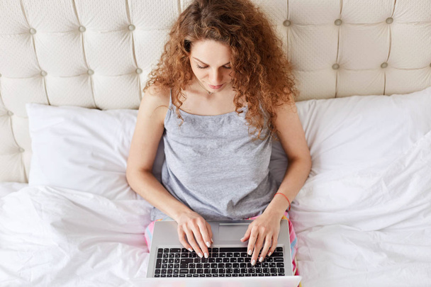 Top view of young woman keyboards information on laptop computer, prepares for passing course paper, sits at bed, being always in touch. Attractive young female chats in social networks online - 写真・画像