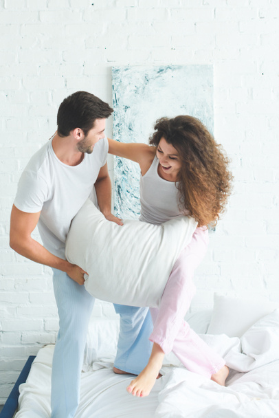 happy young couple in pajamas fighting with pillow on bed - Foto, Imagen