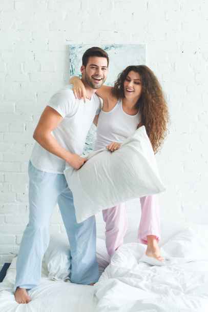 onnellinen nuori pari pyjamassa tilalla tyyny ja seisoo sängyllä
 - Valokuva, kuva