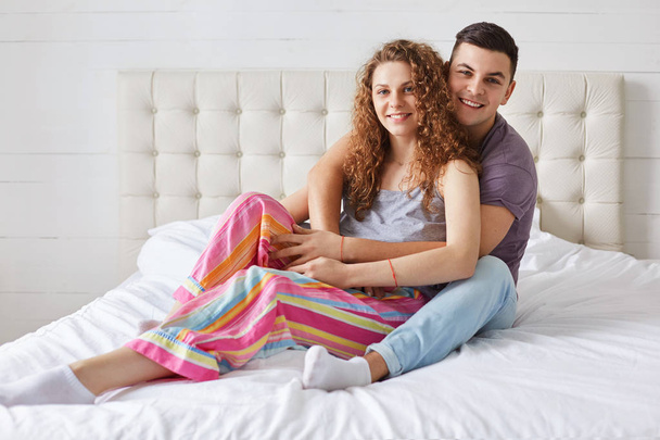 Young family couple sit and embrace on comfortable bed, enjoy togethernesss and good morning, have positive expressions, wear pyjamas. Curly beautiful woman spends free time in bedroom with lover - Foto, afbeelding
