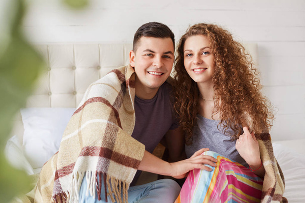 Cheerful girlfriend and boyfriend dressed casually, enjoy coziness under warm plaid in bedroom, have positive expressions, embrace each other and have pleasant talk. Romantic couple in love. - Photo, Image