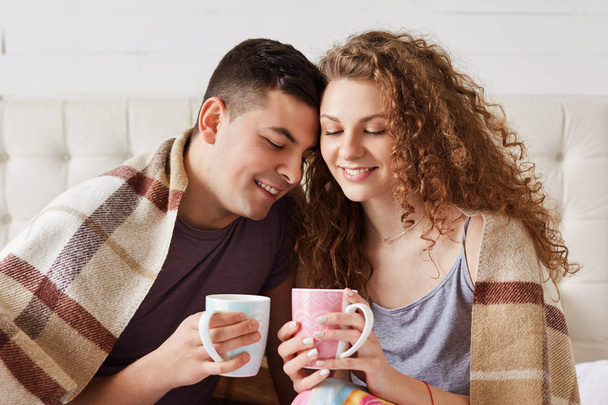 Horizontal shot of beautiful young female and her boyfriend enjoy morning coffee on bed, try to warm themselves after walk on frosty weather, sit close to each other, feel relaxation and support - Photo, image
