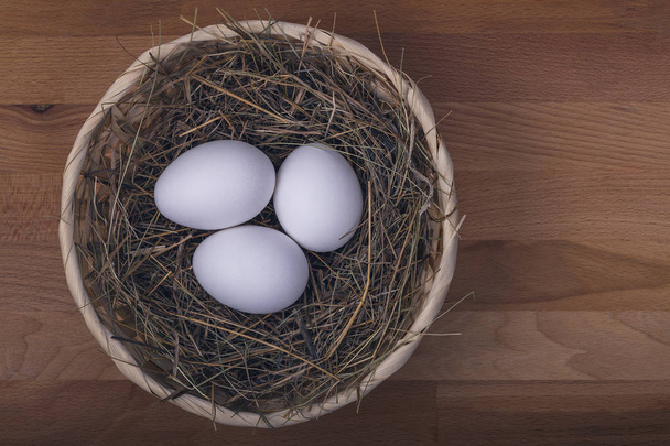 three white chicken eggs in a basket with straw on a wooden background - Φωτογραφία, εικόνα