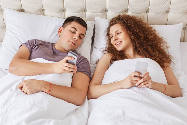 Happy affectionate couple spend morning time in bedroom, being always in touch, use modern gadgets for surfing internet and online communication. Family with mobile phones in comfortable bed - Φωτογραφία, εικόνα