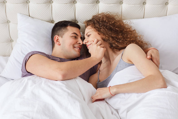 Affectionate couple embrace passionaltely in bed, look at each other`s eyes, feel relaxed, demosntrate mutual love and perfect relationships. Lovely young boyfriend and girlfriend in bedroom. - Foto, Imagem