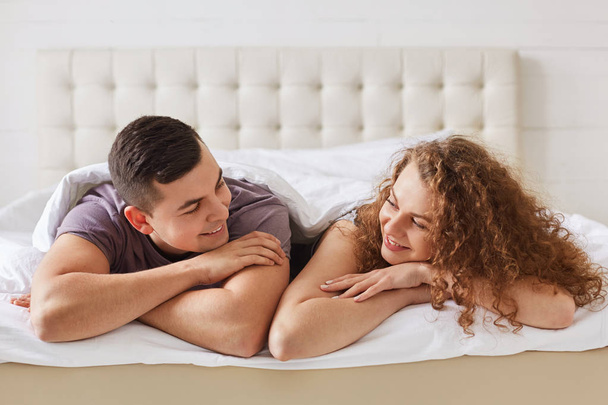 Positive young female and male lie on stomach in comfortable bed, look happily at each other, enjoy good time in bedroom. Romantic couple smile joyfully, enjoy good morning. Relaxation concept - Fotoğraf, Görsel