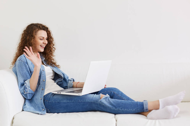 Relaxed positive fashionable female wears denim shirt and jeans, makes video call, happy to see best friend, waves with hand in camera of laptop computer, uses free high speed internet at home - Φωτογραφία, εικόνα