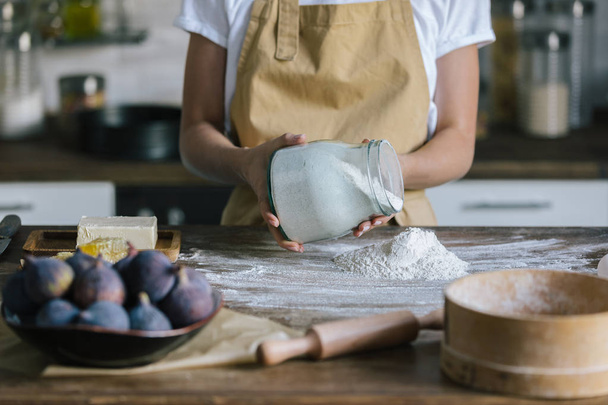 cropped shot of woman pouring flour on rustic wooden table with fig pie ingredients - Photo, image