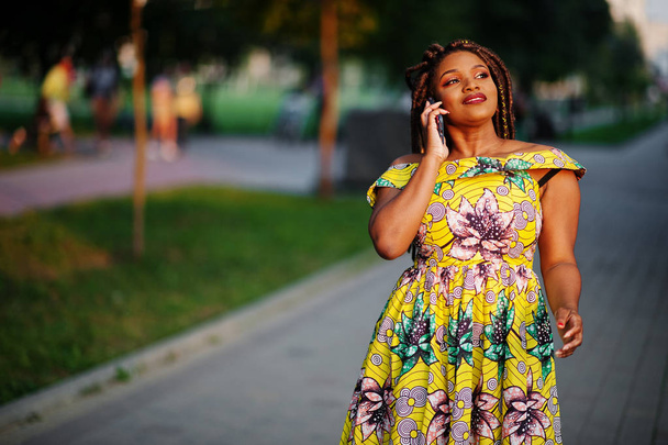 Cute small height african american girl with dreadlocks, wear at coloured yellow dress, posed at sunset, walking and speaking on cellphone. - Zdjęcie, obraz