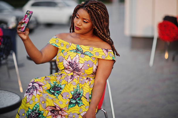 Cute small height african american girl with dreadlocks, wear at coloured yellow dress, sitting at outdoor cafe on red chair and making selfie. - Φωτογραφία, εικόνα
