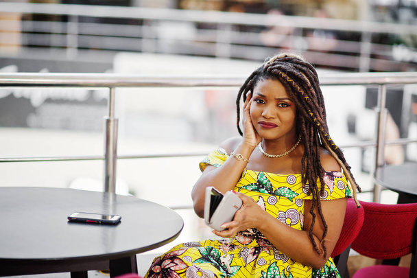 Cute small height african american girl with dreadlocks, wear at coloured yellow dress, sitting on cafe at shopping center and looking at her wallet. No money concept. - Foto, Imagem