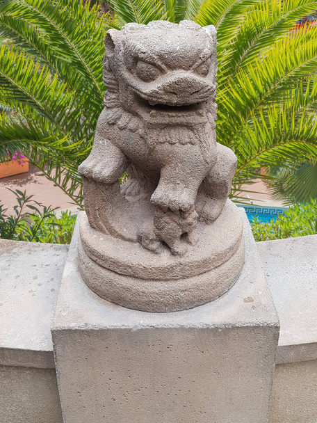 Ancienne statue de lion empereur chinois sur un piédestal en pierre en arrière-plan de beaux palmiers verts une finca avec une piscine
. - Photo, image
