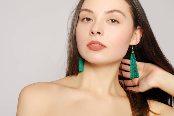 Portrait of young beautiful woman with perfect skin and bright make-up touching her face with manicured fingers. Long green fabric earrings in her ears. Her shoulders naked. Close up. Copy-space - Фото, изображение