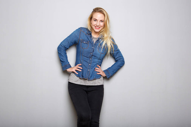 Portrait of young blond woman smiling with hands on hip against gray background - Photo, Image