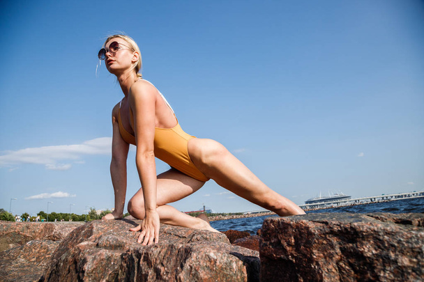 Beauty blond woman wearing sunglasses and swimsuit making fitness on rocky beach at daytime - Photo, Image