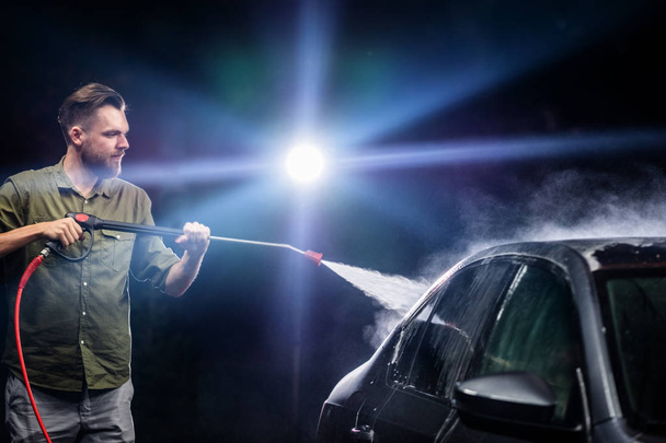 Un hombre con barba o lavadora de coches lava un coche gris con un aparato de alta presión por la noche en un lavado de coches
 - Foto, imagen
