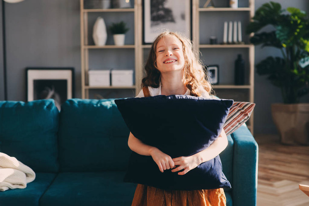 happy cheerful kid girl posing with pillow, cozy couch behind in modern living room. Buying new home, moving to big apartments, house rental concept. - Foto, afbeelding