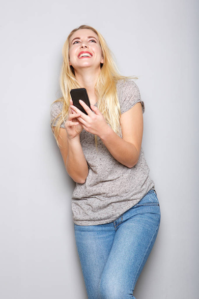 Портрет счастливой молодой блондинки с помощью мобильного телефона за серой стеной
 - Фото, изображение