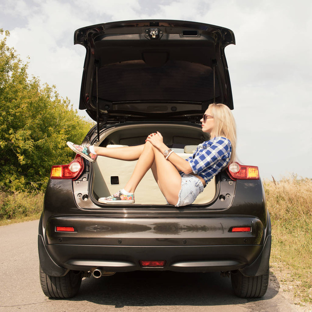 Young attractive woman sitting in the open trunk of a car. Summer road trip - Photo, image