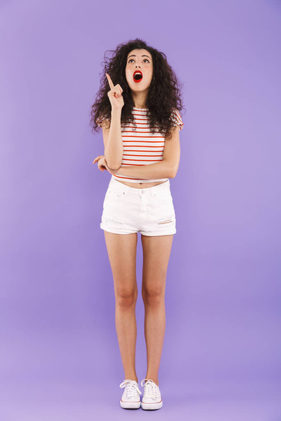 Full length portrait of astonished teenage girl with curly hair in summer wear smiling and looking upward isolated over violet background in studio - 写真・画像
