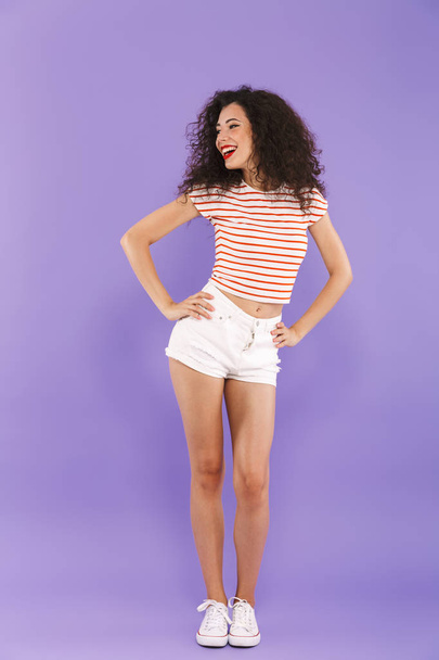 Full length portrait of pretty young woman with curly hair in summer wear smiling and looking aside isolated over violet background in studio - Valokuva, kuva