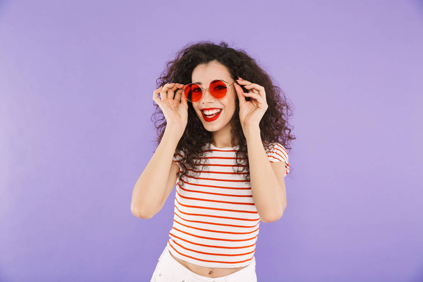 Photo of pretty trendy woman with curly hair in summer wear and sunglasses looking forward isolated over violet background in studio - Foto, Imagem