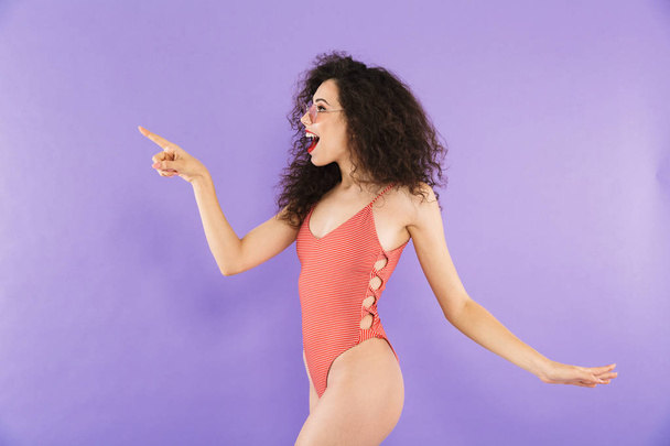 Portrait of a beautiful young woman dressed in swimsuit posing while standing isolated, pointing away - 写真・画像