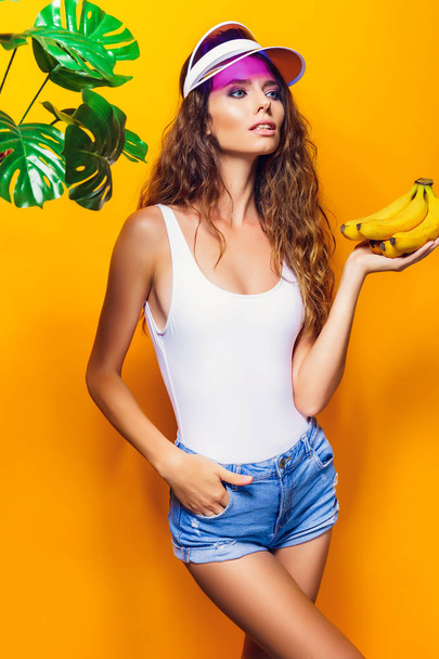Woman in swimsuit and blue shorts holding banana and posing isolated over yellow background - Фото, зображення